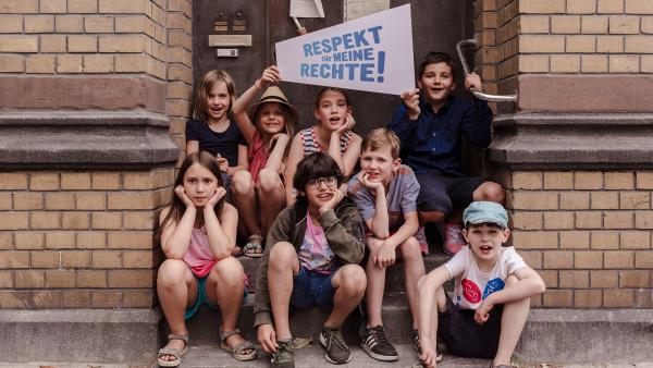Kinder sitzen auf einer Treppe vor einer Haustür und halten ein Schild mit dem Schriftzug: Respekt für meine Rechte! Abenteuer digital.