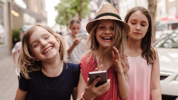 Drei Mädchen laufen lachend eine Straße entlang. Eine von ihnen hat ein Smartphone in der Hand.