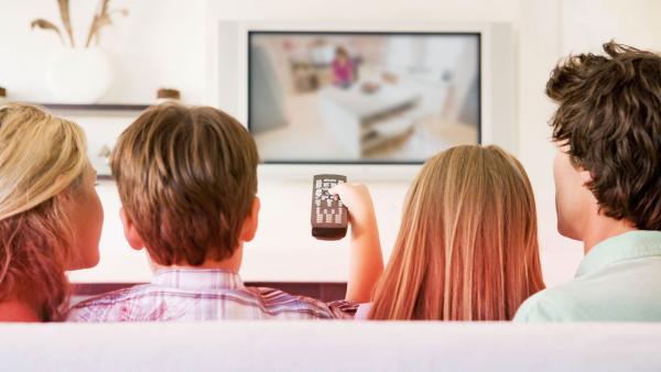 Eine Familie sitzt vor dem Fernseher. Einer hält die Fernbedienung in der Hand.