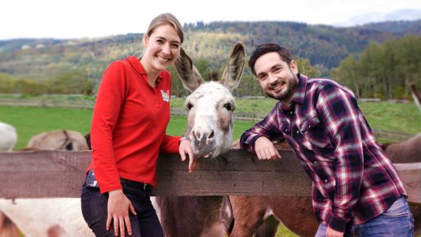 Die Moderator*innen Melanie und Sebastian stehen neben einem Esel und lächeln in die Kamera.