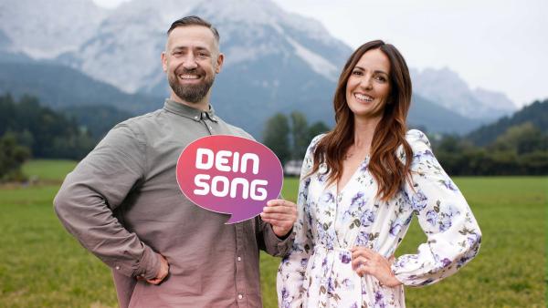 Johanna Klum und Bürger Lars Dietrich stehen in den Bergen und halten das Logo von "Dein Song" in der Hand.