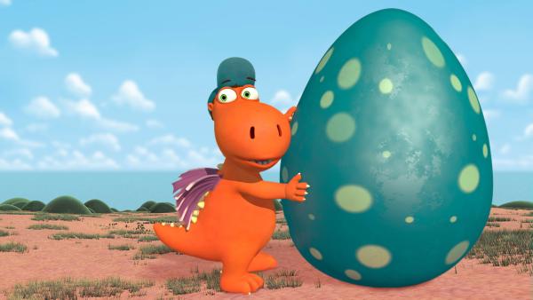 Der Drache Kokosnuss hält ein riesiges Ei fest, das neben ihm steht. Das Ei ist größer als er.