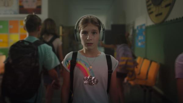 Eileen trägt Kopfhörer, ihren Ranzen und ein T-shirt mit Regenbogenmotiv.