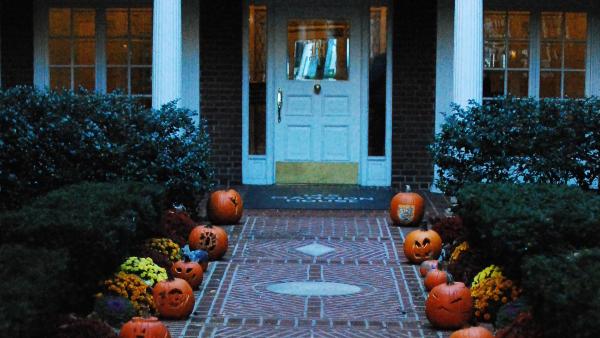 Auf diesem Bild sieht man einen Hauseingang mit Vorgarten. Im Vorgarten sind viele Halloween Kürbisse aufgestellt, in die Gesichter geschnitzt wurden. Der Hintergrund ist dunkel wie bei Anbruch der Nacht.