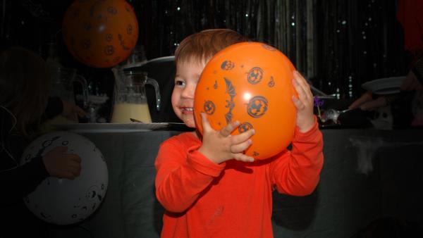 Auf diesem Bild sieht man einen kleinen Jungen, der einen orangefarbenen Luftballon hält. Auf diesem Luftballon sind Halloween-Kürbisse und Fledermäuse abgebildet. Der Junge lacht. Der Hintergrund ist dunkel.