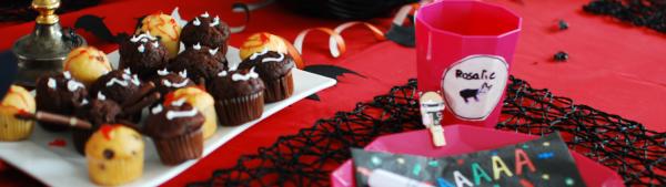 Auf diesem Bild sieht man einen bunt geschmückten Halloween-Tisch mit roter Tischdecke. Auf dem Tisch sieht man einen Teller mit Serviette, einen Becher und einen Teller mit kleinen Muffins. Der Halloweenparty-Tisch ist mit kleinen schwarzen Fledermäusen aus Papier und mit orangefarbenen Luftschlangen dekoriert. Die Tischdecke ist rot. 