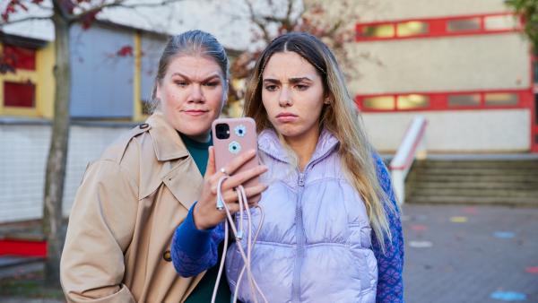 Eine Jugendliche mit langen blonden Haaren schaut auf ein Smartphone, das sie vor sich in der Hand hält. Links neben ihr steht eine erwachsene Frau, die ebenfalls auf das Smartphone schaut.