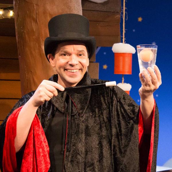 Juri Tetzlaff im KiKA-Baumhaus trägt einen schwarzen Zylinder, einen schwarz-roten Umhang und hält einen Zauberstab sowie ein Glas Wasser mit einem schwimmenden Ei.