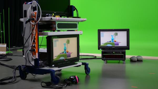 Zwei Bildschirme und weitere technische Geräte stehen in einem Studio. Im Hintergrund ist ein Greenscreen. Auf beiden Bildschrimen sieht man Kikaninchen.