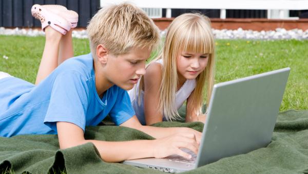 Ein Junge mit kurzen blonden Haaren und ein Mädchen mit langen blonden Haaren liegen auf einer Decke auf einer Wiese. Vor ihnen steht ein aufgeklappter Laptop.