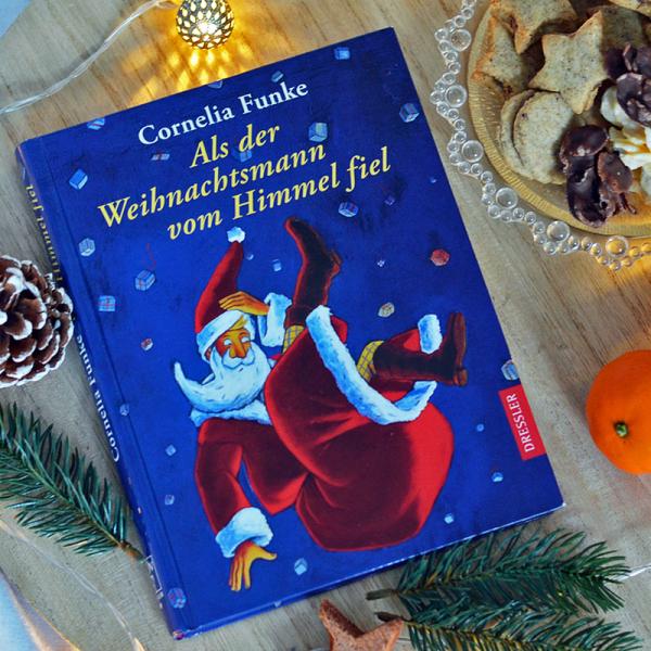 Das Buch "Als der Weihnachtsmann vom Himmel fiel" von Cornelia Funke liegt auf einem Holzbrett. Es ist blau mit gelber Schrift und ein Weihnachtsmann ist darauf zu sehen. Rund herum liegen Tannenzweige und Zapfen, sowie eine Lichterkette. Eine Schale mit Plätzchen und zwei Mandarinen liegen rechts daneben.
