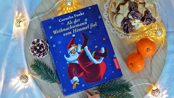 Das Buch "Als der Weihnachtsmann vom Himmel fiel" von Cornelia Funke liegt auf einem Holzbrett. Es ist blau mit gelber Schrift und ein Weihnachtsmann ist darauf zu sehen. Rund herum liegen Tannenzweige und Zapfen, sowie eine Lichterkette. Eine Schale mit Plätzchen und zwei Mandarinen liegen rechts daneben.