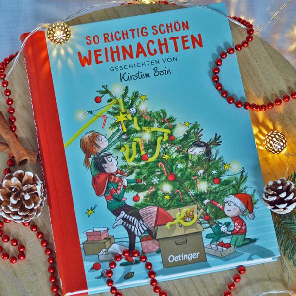 Das Buch "So richtig schön Weihnachten" von Kirsten Boie liegt auf einem Holzbrett. Es ist blau mit roter Schrift und auf dem Buchcover schmücken Kinder zusammen mit ihrem Papa einen Weihnachtsbaum. Rund um das Buch liegen Tannenzweige und Zapfen, sowie eine Lichterkette. Eine rot glänzende Perlenkette sowie zwei Zimtstangen sind um das Buch gelegt.