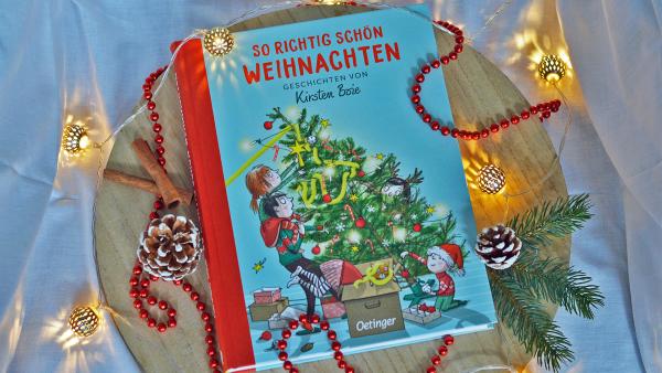 Das Buch "So richtig schön Weihnachten" von Kirsten Boie liegt auf einem Holzbrett. Es ist blau mit roter Schrift und auf dem Buchcover schmücken Kinder zusammen mit ihrem Papa einen Weihnachtsbaum. Rund um das Buch liegen Tannenzweige und Zapfen, sowie eine Lichterkette. Eine rot glänzende Perlenkette sowie zwei Zimtstangen sind um das Buch gelegt.
