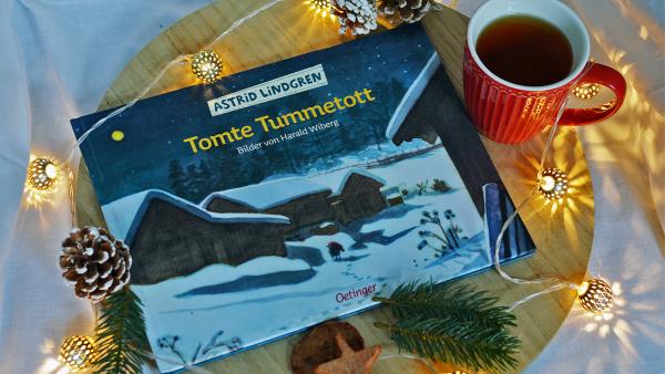 Das Buch Tomte Tummetott von Astrid Lindgren liegt auf einem Holzbrett. Rund herum liegen Tannenzweige und Zapfen, sowie eine Lichterkette. Eine rote Teetasse steht rechts daneben.