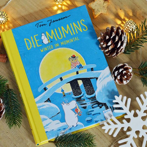 Das Buch "Die Mumins- Winter im Mumintal" von Tove Jansson liegt auf einem Holzbrett. Es ist blau mit gelber Schrift und der nilpferdartige Mumintroll ist darauf zu sehen. Um das Buch herum liegen Tannenzweige und Zapfen, sowie eine Lichterkette. Eine große gebastelte Schneeflocke liegt rechts unten daneben.