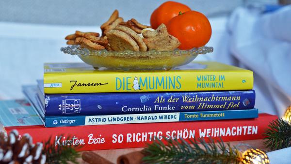 4 Bücher liegen übereinander gestapelt auf einem Holzbrett. So richtig schön Weihnachten von Kirsten Boie, Tomte Tummetott von Astrid Lindgren, Als der Weihnachtsmann vom Himmel fiel von Cornelia Funke und Die Mumins- Winter im Mumintal von Tove Jansson. Auf dem Bücherstapel steht eine Schale mit Plätzchen und Mandarinen. Vor dem Bücherstapel liegen Tannenzweige und Zapfen sowie eine Lichterkette.