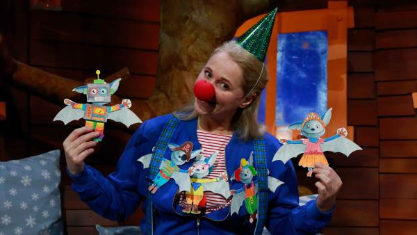 Eine blonde Frau mit einer blauen Jacke, einem grünen Partyhut und einer roten Clownsnase lächelt und hält Bastelbögen in den Händen.