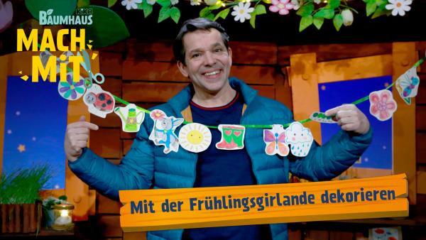Ein fröhlicher Mann in blauer Jacke hält eine selbstgebastelte Frühlingsgirlande mit bunten Motiven aus Blumen, Schmetterlingen und Käfern. Im Hintergrund ist eine gemütliche Holzdeko mit Pflanzen und Lichterketten zu sehen. Der Text „Mit der Frühlingsgirlande dekorieren“ ist auf einem orangefarbenen Banner zu lesen.