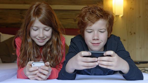 Ein Mädchen und ein Junge liegen auf dem Bett und schauen jeweils auf ihr Smartphone.