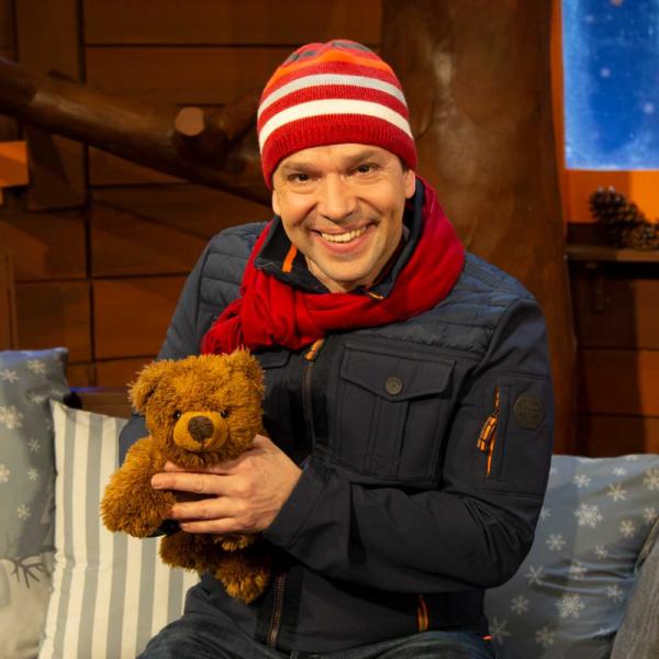 Juri Tetzlaff im KiKA-Baumhaus trägt eine rot-weiße Mütze, einen roten Schal und eine Jacke und hält einen braunen Teddybär in den Händen.