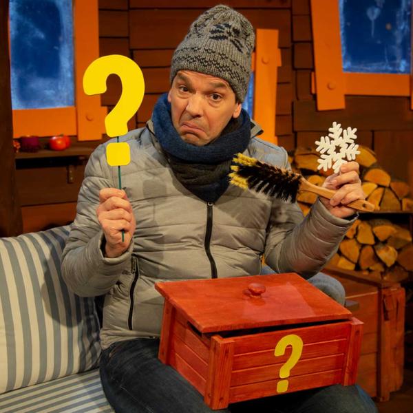Juri Tetzlaff im KiKA-Baumhaus trägt eine Mütze, Schal und Jacke. Er hält ein gelbes Fragezeichen, einen Feger, ein Eiskristall und auf seinem Schoss liegt eine geöffnete, rote Kiste mit einem gelben Fragezeichen auf der Vorderseite.
