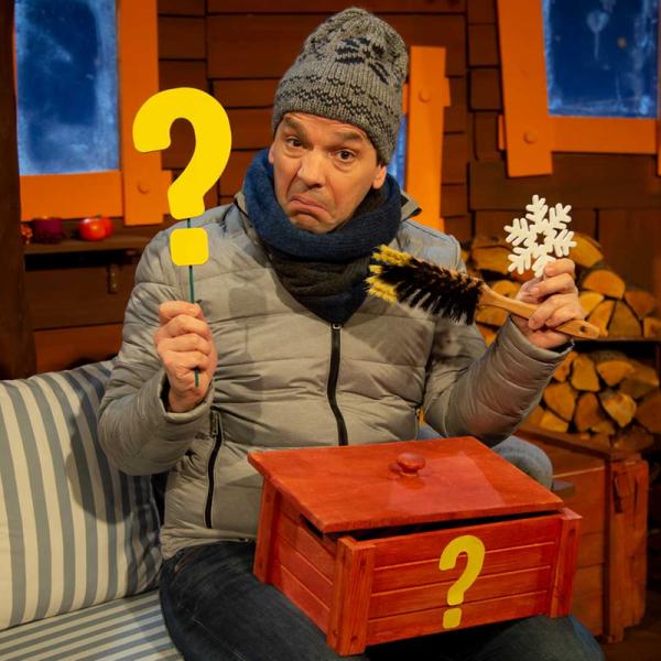 Juri Tetzlaff im KiKA-Baumhaus trägt eine Mütze, Schal und Jacke. Er hält ein gelbes Fragezeichen, einen Feger, ein Eiskristall und auf seinem Schoss liegt eine geöffnete, rote Kiste mit einem gelben Fragezeichen auf der Vorderseite.