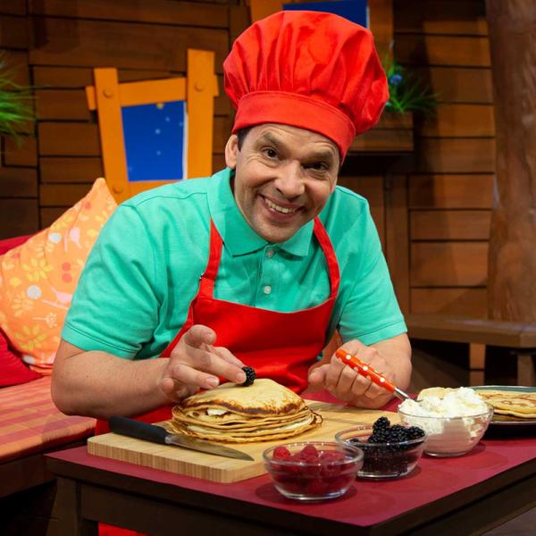 Juri Tetzlaff im KiKA-Baumhaus garniert Pfannkuchen mit Beeren und trägt eine rote Kochschürze sowie eine Kochmütze. 
