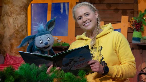 Eine blone Frau in gelbem Pullover (Singa) hält ein aufgeschlagenes Buch in der Hand. Links neben ihr sitzt eine blaue Feldermaus-Puppe (Fidi) schaut in das aufgeschlagene Buch.