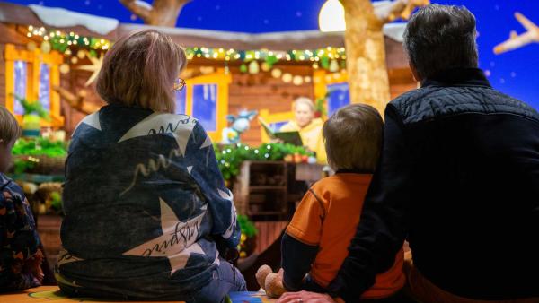 Im Vordergrund sind eine Frau mit schulterlangen blonden Haaren, ein Kind mit kurzen dunkelblonden Haaren und ein Mann mit dunklen Haaren von hinten zu sehen. Unscharf im Hintergrund ist das Set des KiKA-Baumhaus zu sehen, in dem Singa und Fidi in ein Buch schauen.