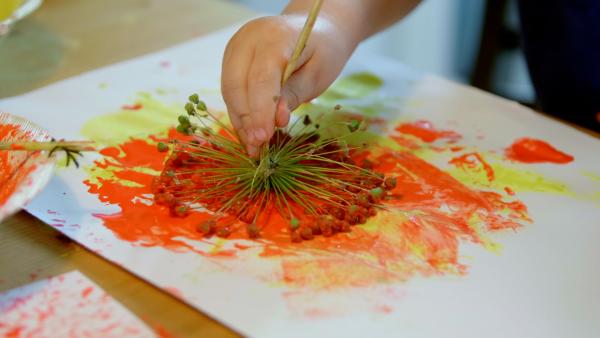 Blume wird mit Farbe auf ein Papier gedruckt. Auf dem Papier entsteht ein orangefarbener Abdruck. 