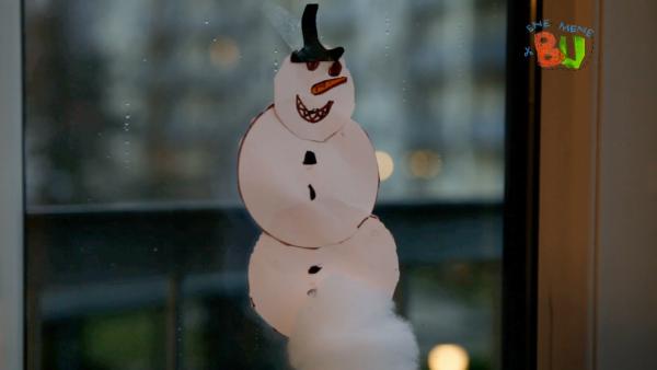 Auf dem Bild sieht man einen selbstgebastelten Schneemann aus Papier, der mit Klebeband auf das Fenster geklebt worden ist.