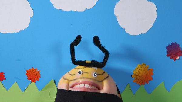 Kinngesicht in Form einer Biene vor einer gebastelten Wiese als Hintergrund. 