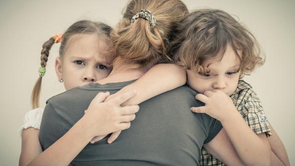 Zwei Kinder, ein Junge und ein Mädchen, umarmen eine Frau und lassen sich trösen.