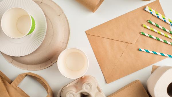 Es liegen diverse Bastelsachen auf einem Tisch. Dazu gehören ein Briefumschlag, Strohhalme, mehrere Pappbecher, ein Pappteller, ein Eierkarton sowie Kolpapier und eine Klopapierrolle