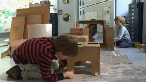 Drei Kinder bauen aus alten Pappkartons eine kleine Wohnung nach mit einem Herd und einem Kühlschrank.
