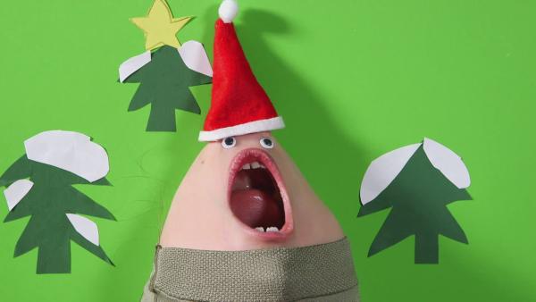 Kinngesicht in Form eines Nikolauses. Gesicht öffnet den Mund weit und ist vor einem grünen gebastelten Hintergrund mit drei Tannen. 