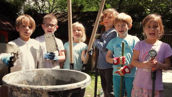 Sechs Kinder halten ihre Werkzeuge, wie Maurerkelle Spaten und einen Wassserschlauch hoch. Sie wollen einen Hühnerstall bauen.