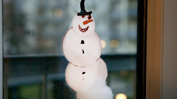 Ein Papierschneemann aus drei Kreisen am Fenster. Der Schneemann hat ein freundliches gemaltes Gesicht. 