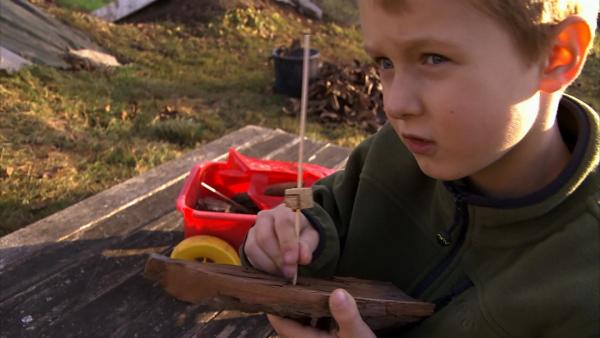 Ein Junge steckt einen Holzstab in ein Stück Rinde. Er sitzt draußen in der Natur.