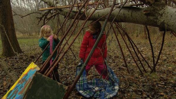 Im Wald bindet ein Mädchen ein Band an einen Ast. Ein anderes Mädchen hält eine Decke in den Händen.