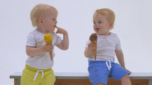Zwei Jungs sitzen auf einer Holzbank. Sie essen Eis und schauen sich dabei an.