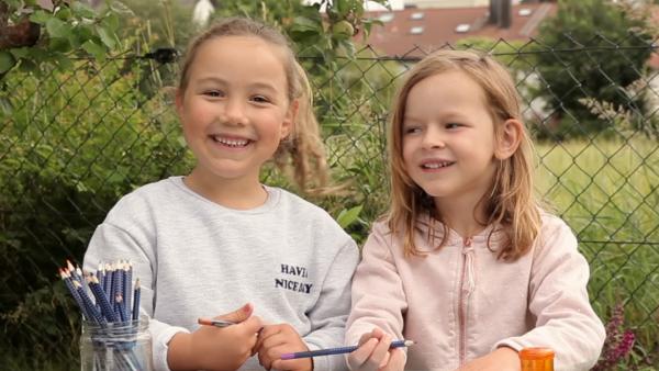 Zwei Mädchen sitzen draußen und halten Stifte in der Hand. Vor ihnen liegt ein Blatt Papier.