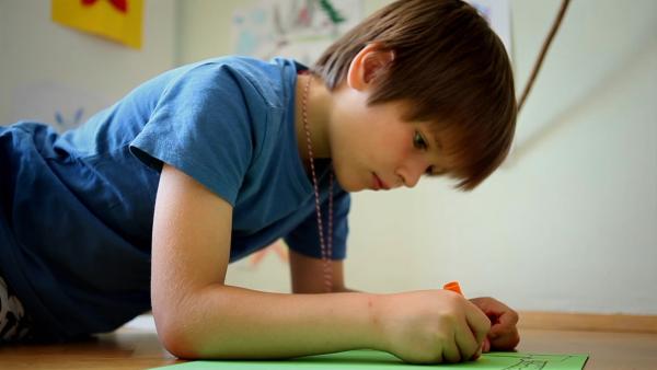 Ein Vorschulkind liegt auf dem Boden und malt mit einem orangenen Stift auf ein grünes Blatt.