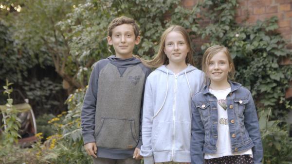 Ein Junge und zwei Mädchen stehen nebeneinander im Garten und blicken lächelnd in die Kamera.