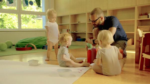 Drei Kleinkinder sitzen vor einem großen Blatt auf dem Boden. Vor ihnen hockt ein Erzieher.