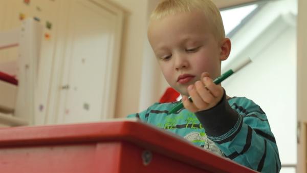 Ein Junge sitzt an einem roten Tisch und malt mit einem grünen Filzstift.