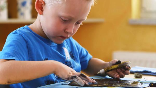 Ein Kind sitzt an einem Tisch und malt mit einem Glitzerstift auf einem blauen Papier.
