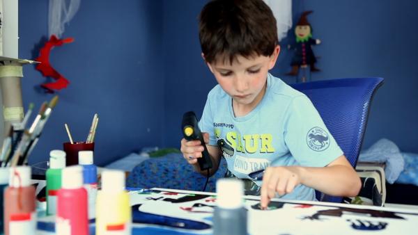 Ein Junge sitzt in seinem blauen Kinderzimmer am Tisch und hält eine Heißklebepistole in der Hand.