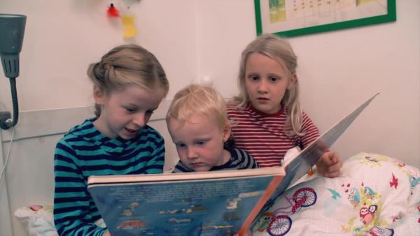 Drei Kinder sitzen auf einem Bett und schauen gemeinsam in ein großes Buch.